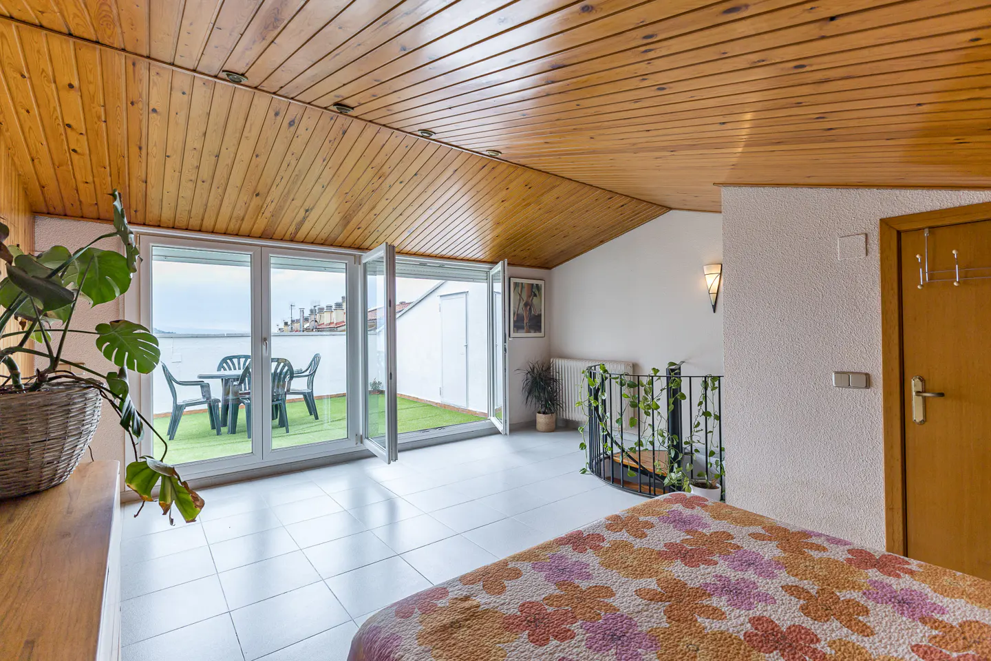 Attic bedroom with wood-paneled ceiling, floral bedspread, and glass doors opening to a balcony with outdoor furniture.