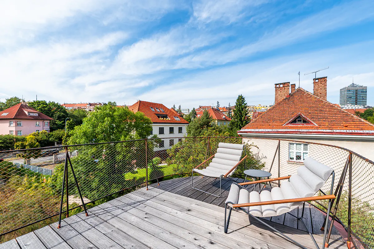 Rooftop patio with two lounge chairs and a small table, overlooking a neighborhood with red-tiled roofs and green trees under a blue sky.
