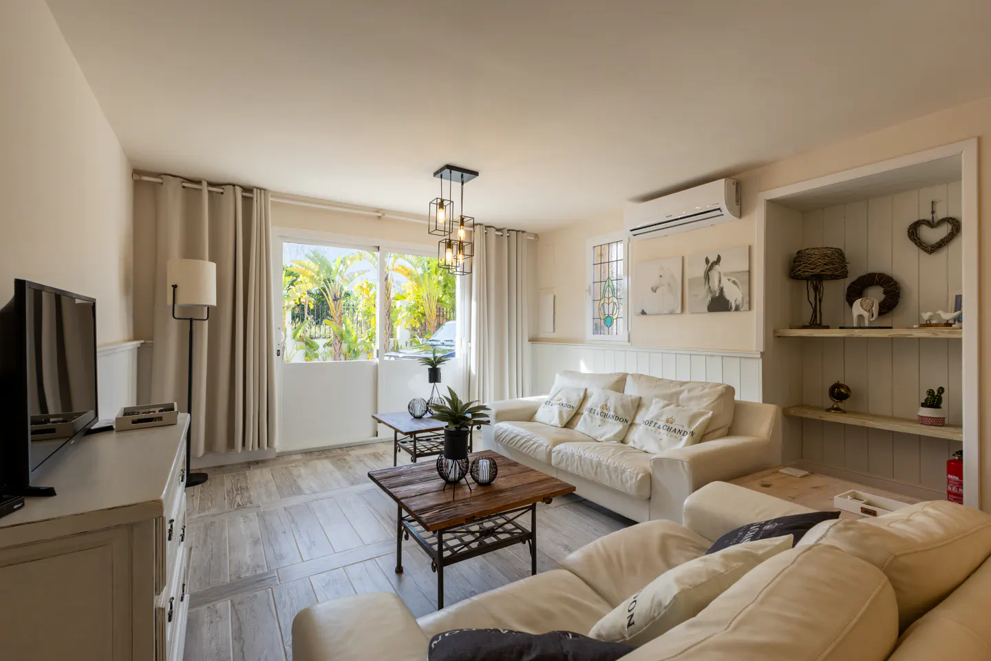 Bright living room with white sofas, wood tables, and a large window showing palm trees outside. Shelves display decorative items.