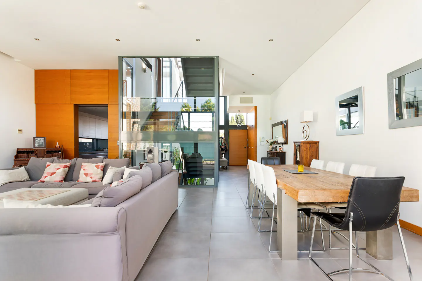 Open-concept living space with a gray sofa, wood dining table with white chairs, and a glass-enclosed staircase.