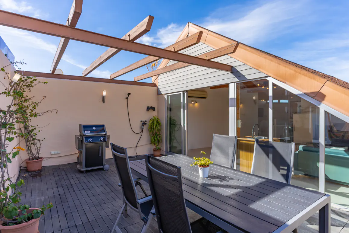 Outdoor rooftop terrace with a dining table, grill, potted plants, and a wooden pergola under a bright blue sky.