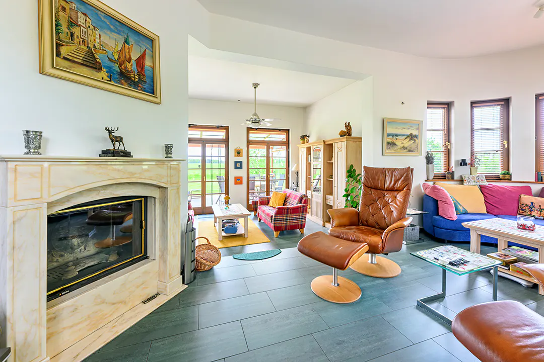 Bright living room with a marble fireplace, leather chair, blue sofa, and a painting of boats on the wall.