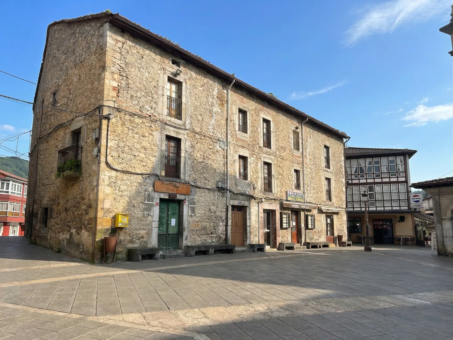 A three-story stone building with multiple windows and doors faces a paved square under a blue sky.