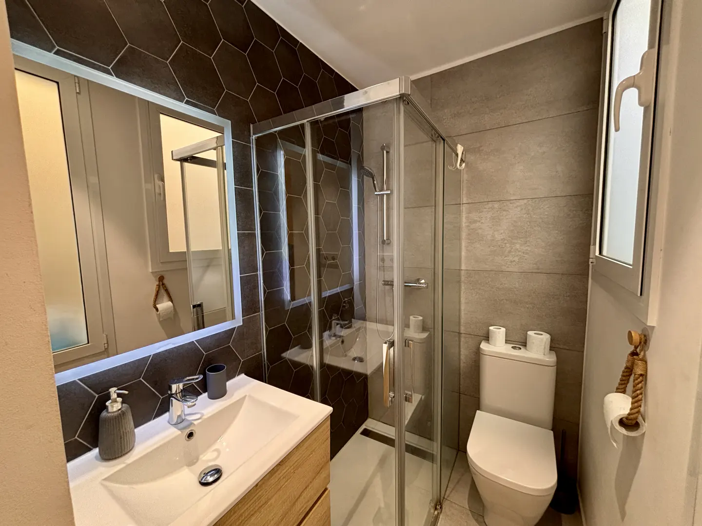 Modern bathroom with a wood vanity, white sink, glass shower stall, toilet, and dark hexagonal tile accent wall.