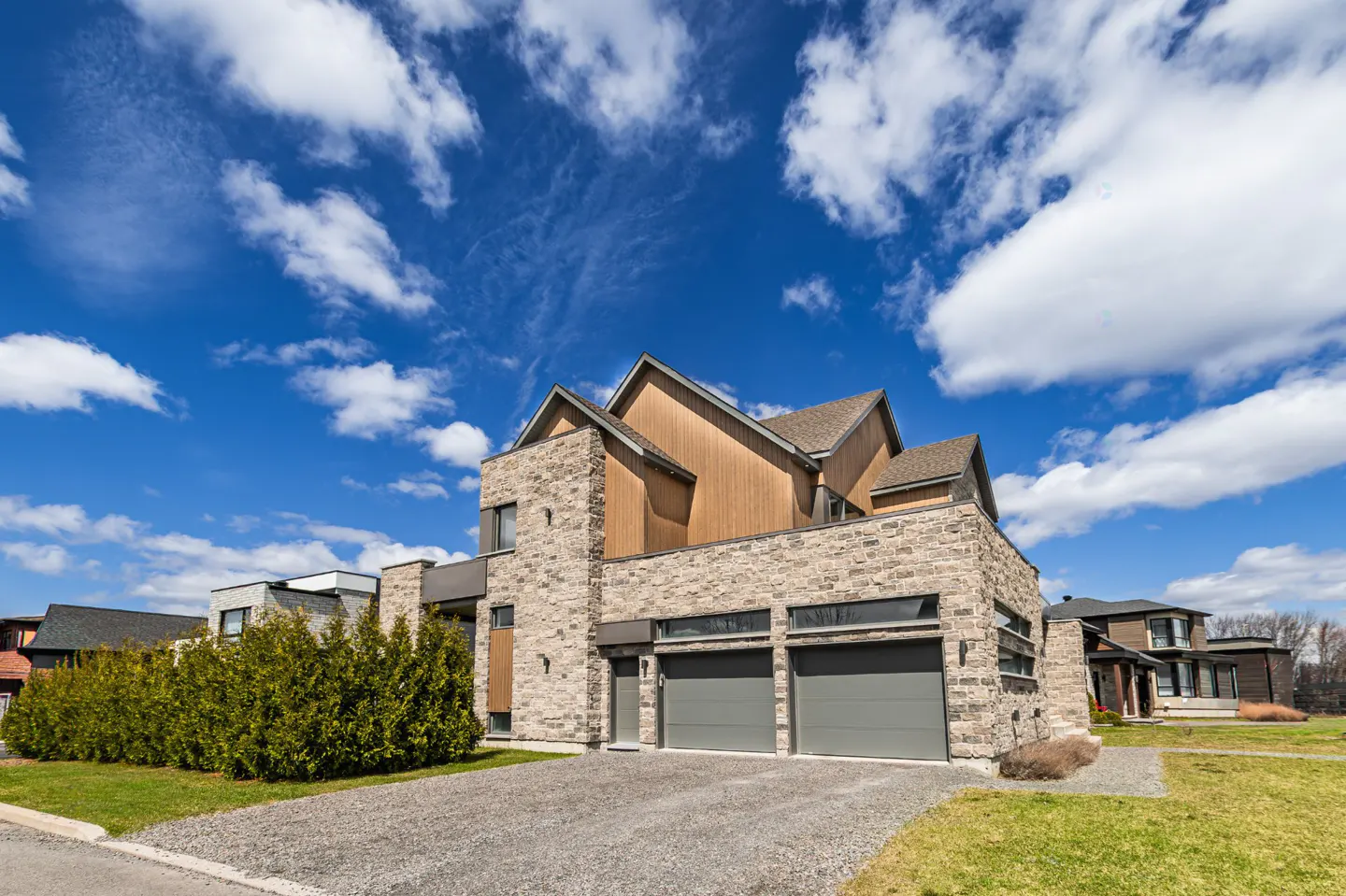 Two-story house with stone and wood facade, gray garage doors, and gravel driveway under a blue sky with white clouds.