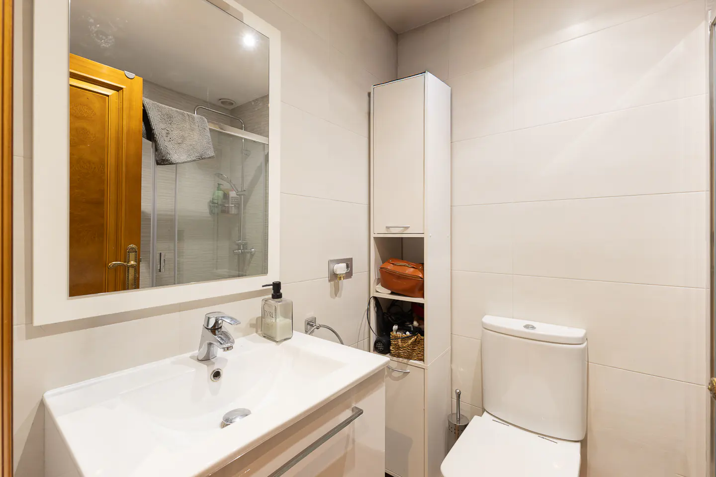 Modern bathroom with white vanity, sink, mirror, tall storage cabinet, and toilet against light-colored tiled walls.