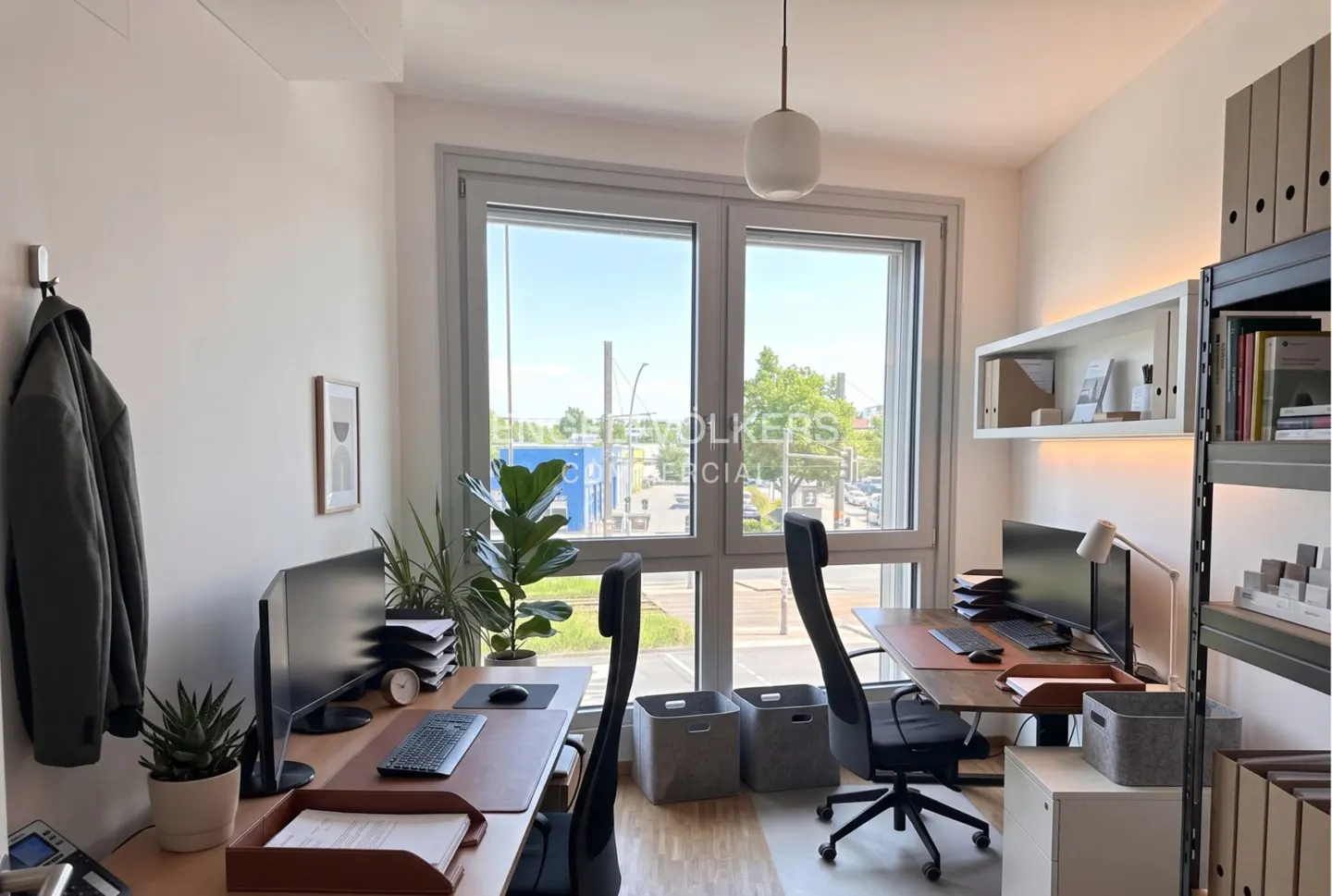 Bright office space with two desks, computers, plants, and a large window with a city view. A coat hangs on the wall.