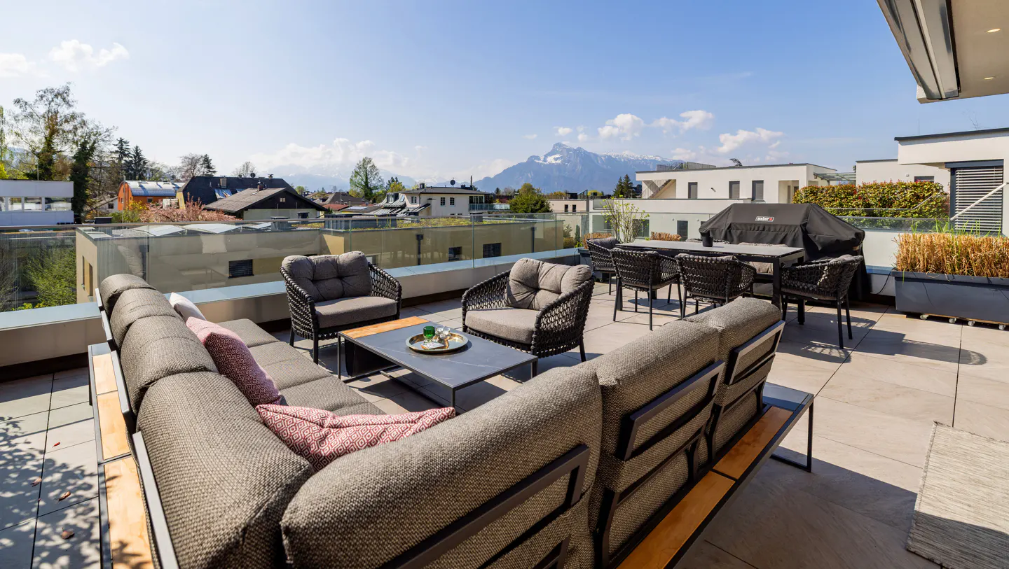 Outdoor patio with gray wicker furniture, a grill, and a mountain view on a sunny day.