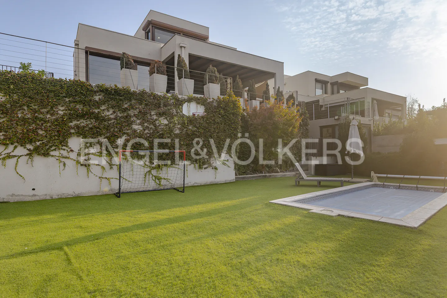 Modern home with a pool and green lawn. A soccer goal sits near a vine-covered wall. A lounge chair and umbrella are near the covered pool.