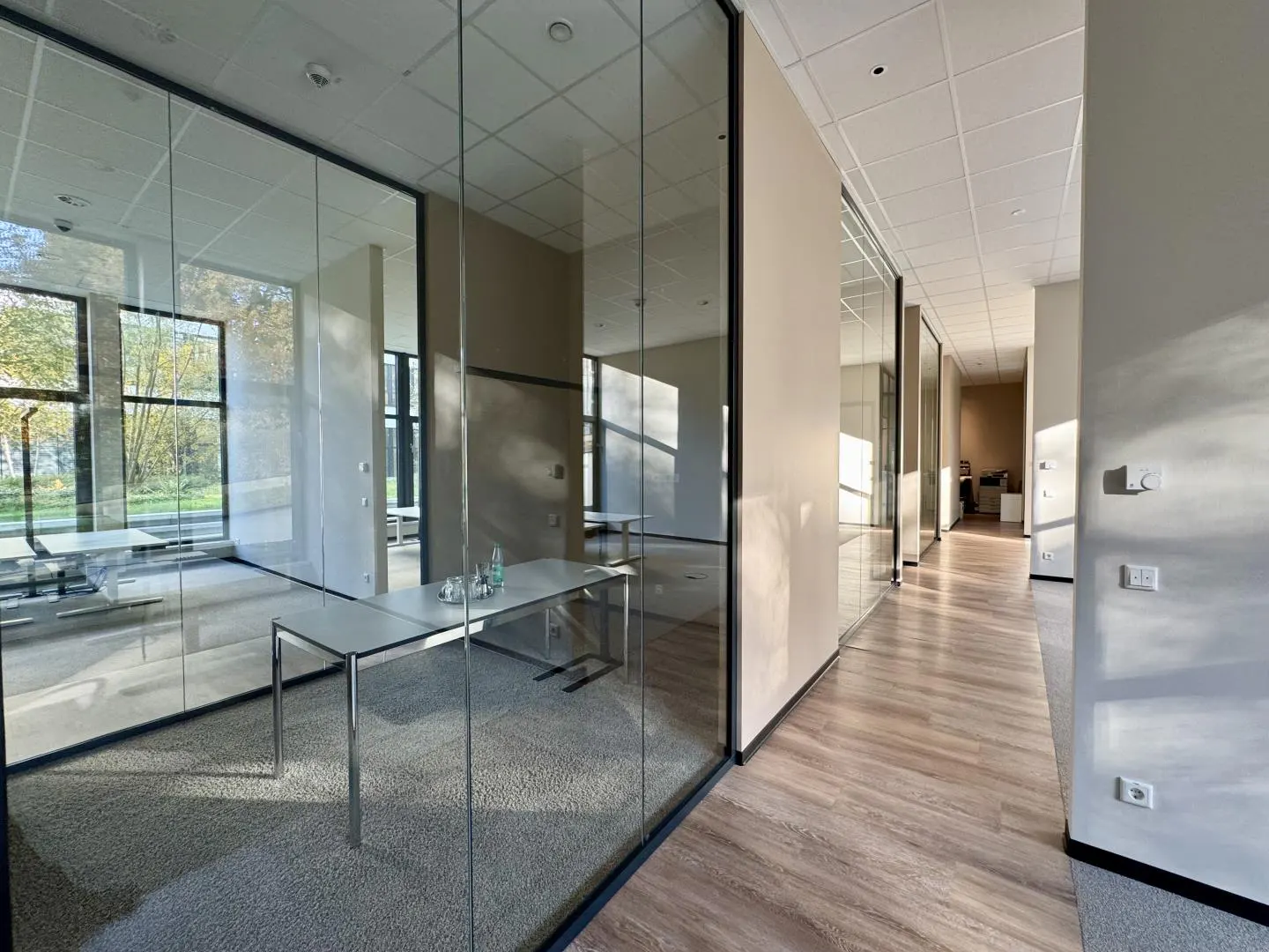 Office space with glass-walled rooms, light wood floors, and a long hallway. Tables and chairs are visible inside the rooms.