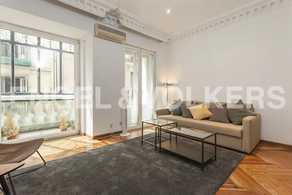 Bright living room with a beige sofa, glass tables, and a large window. A gray rug covers part of the parquet floor.