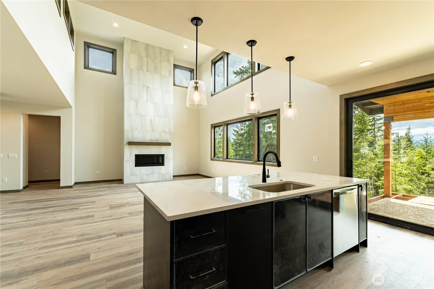 Bright, modern kitchen with a black island, white countertops, and pendant lights. Large windows offer a view of lush greenery.
