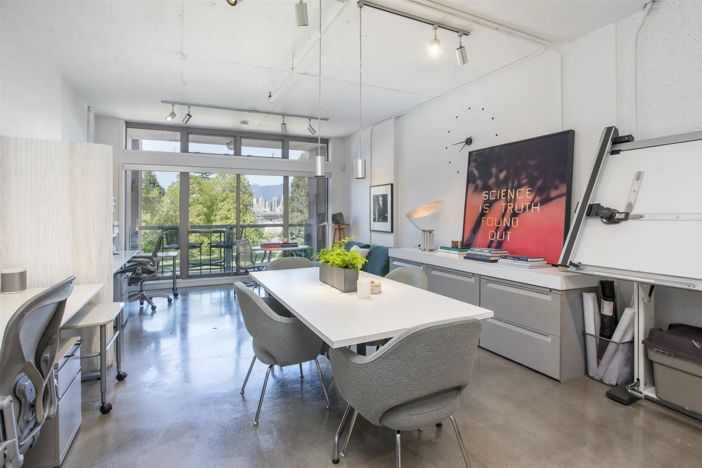Bright, modern office space with a white table surrounded by gray chairs. Large windows offer a view of trees and a cityscape. Artwork and a drafting table add character.