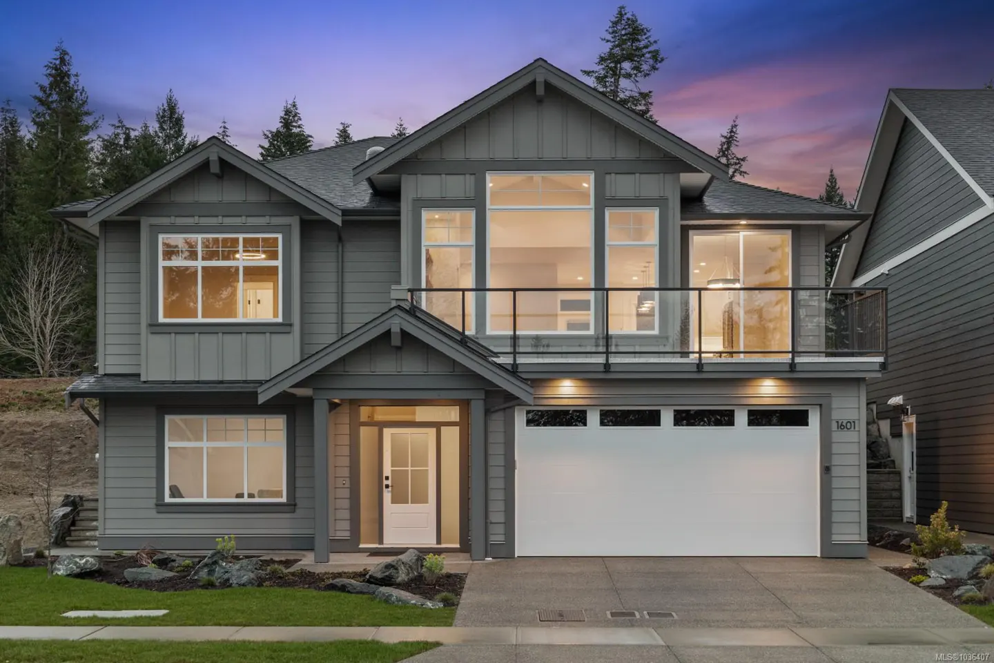 Two-story gray house with white trim, a white garage door, and a glass-railed balcony. Trees are in the background.