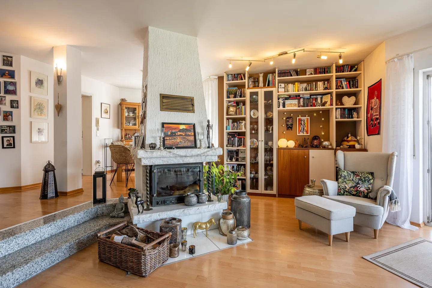 Bright living room with a stone fireplace, hardwood floors, and a large bookshelf. A cozy armchair and ottoman sit near a window.