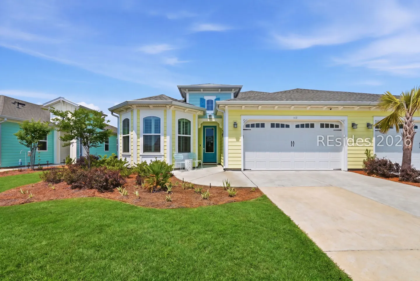 A yellow house with a teal door and white garage door sits on a green lawn under a blue sky.