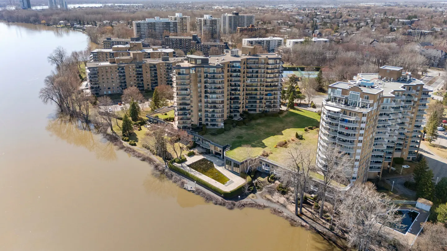 Aerial view of tan apartment buildings with balconies along a riverbank, trees, and city skyline in the background.