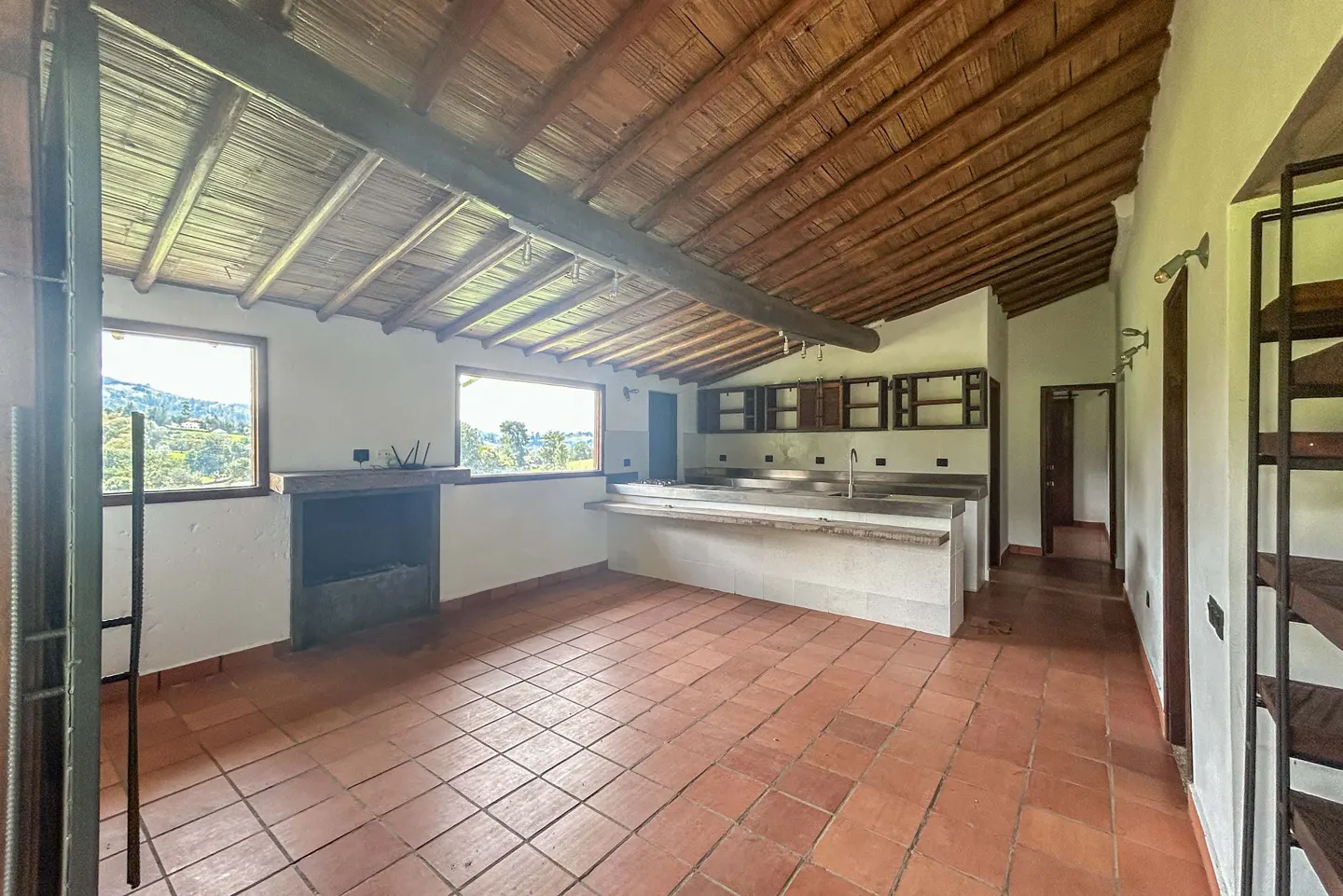Open-plan kitchen with terracotta tile floor, white walls, and a wooden ceiling. Windows offer a view of green hills.