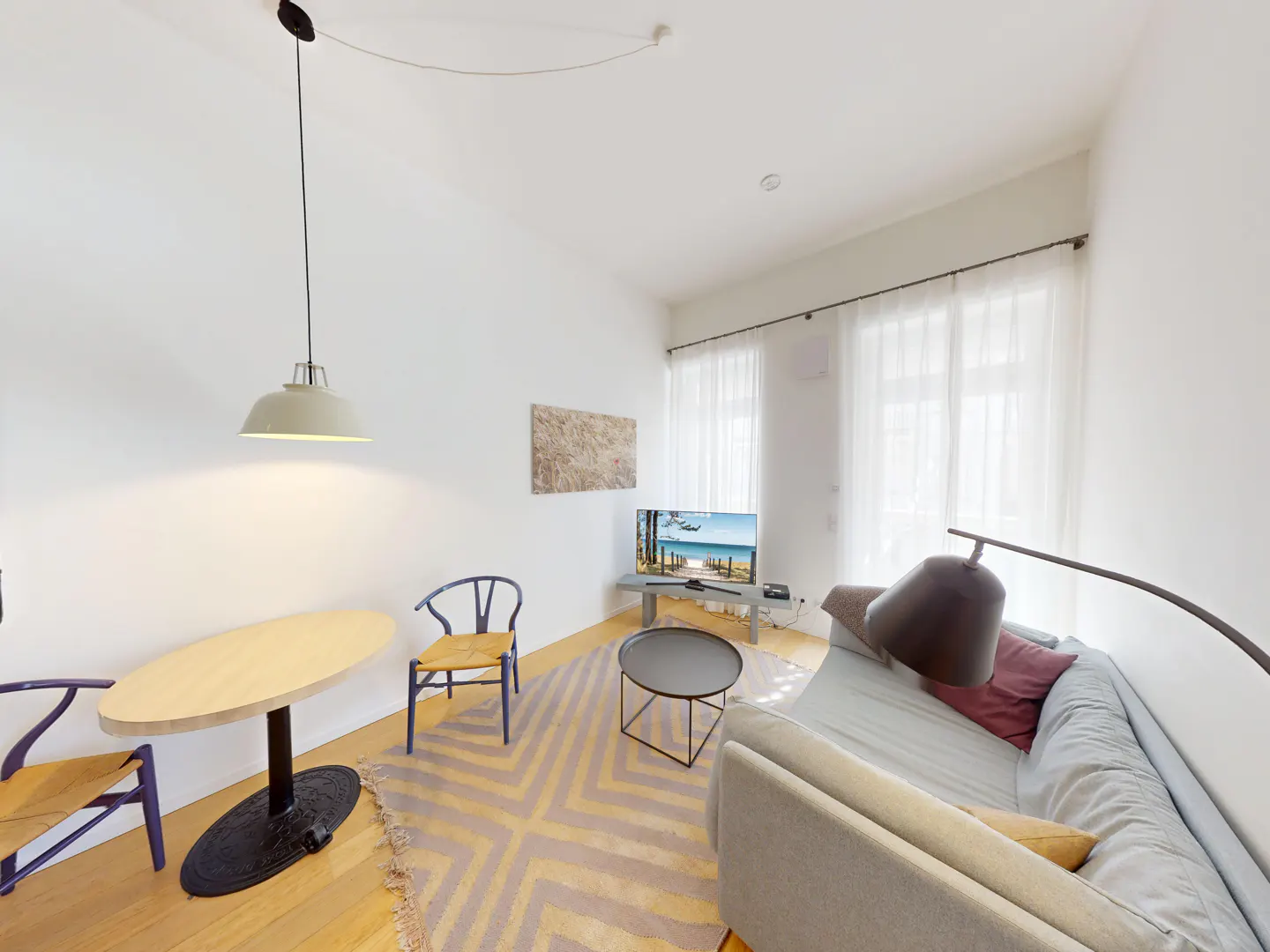 Bright living room with white walls, wood floors, and a geometric rug. A gray sofa faces a TV showing a beach scene. A round table and blue chairs sit under a pendant light.