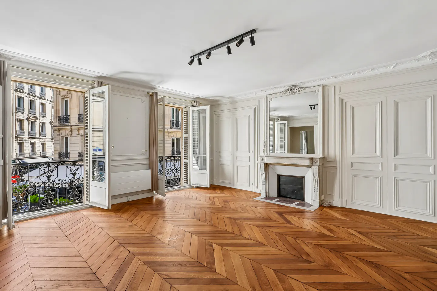 Bright, empty room with herringbone wood floors, white walls, and open French doors to balconies with city views. A fireplace with a mirror above it is on the right.