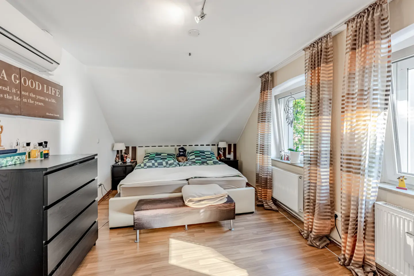 A bright bedroom with a bed, bench, and dark dresser. Windows with striped curtains let in sunlight. The room has a sloped ceiling.