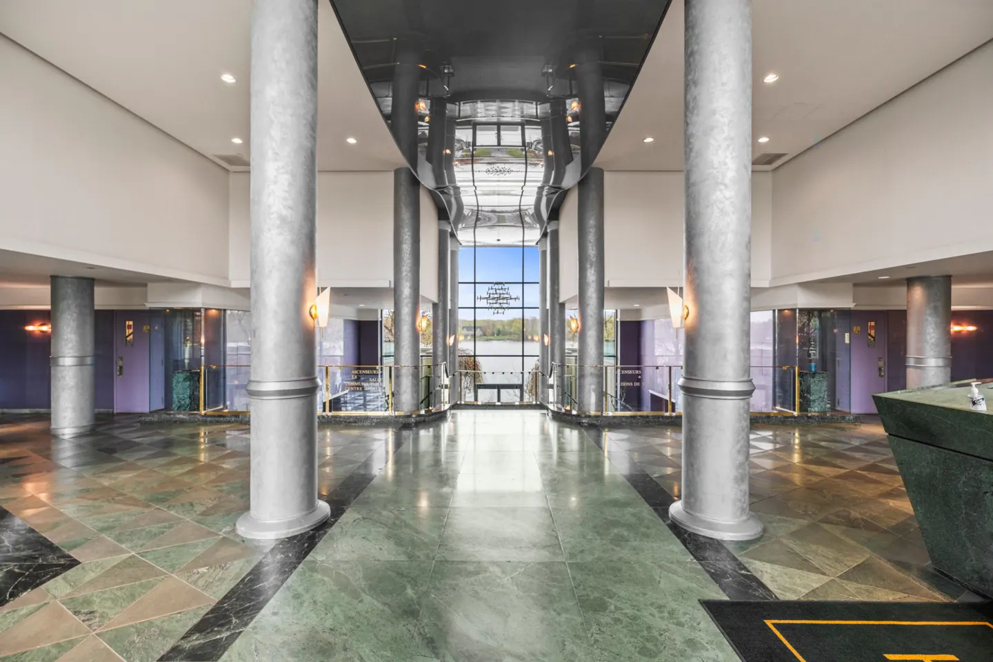 Lobby with marble floors, silver columns, and a view of a lake through a large window. Elevators are on the left.