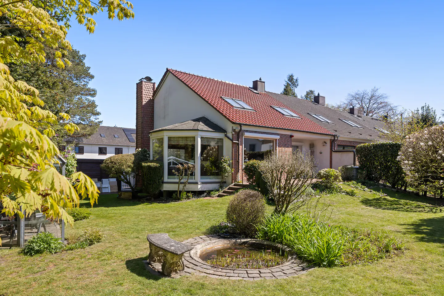Backyard view of a house with a red tile roof, a brick chimney, and a sunroom. A small pond and bench sit on the green lawn.