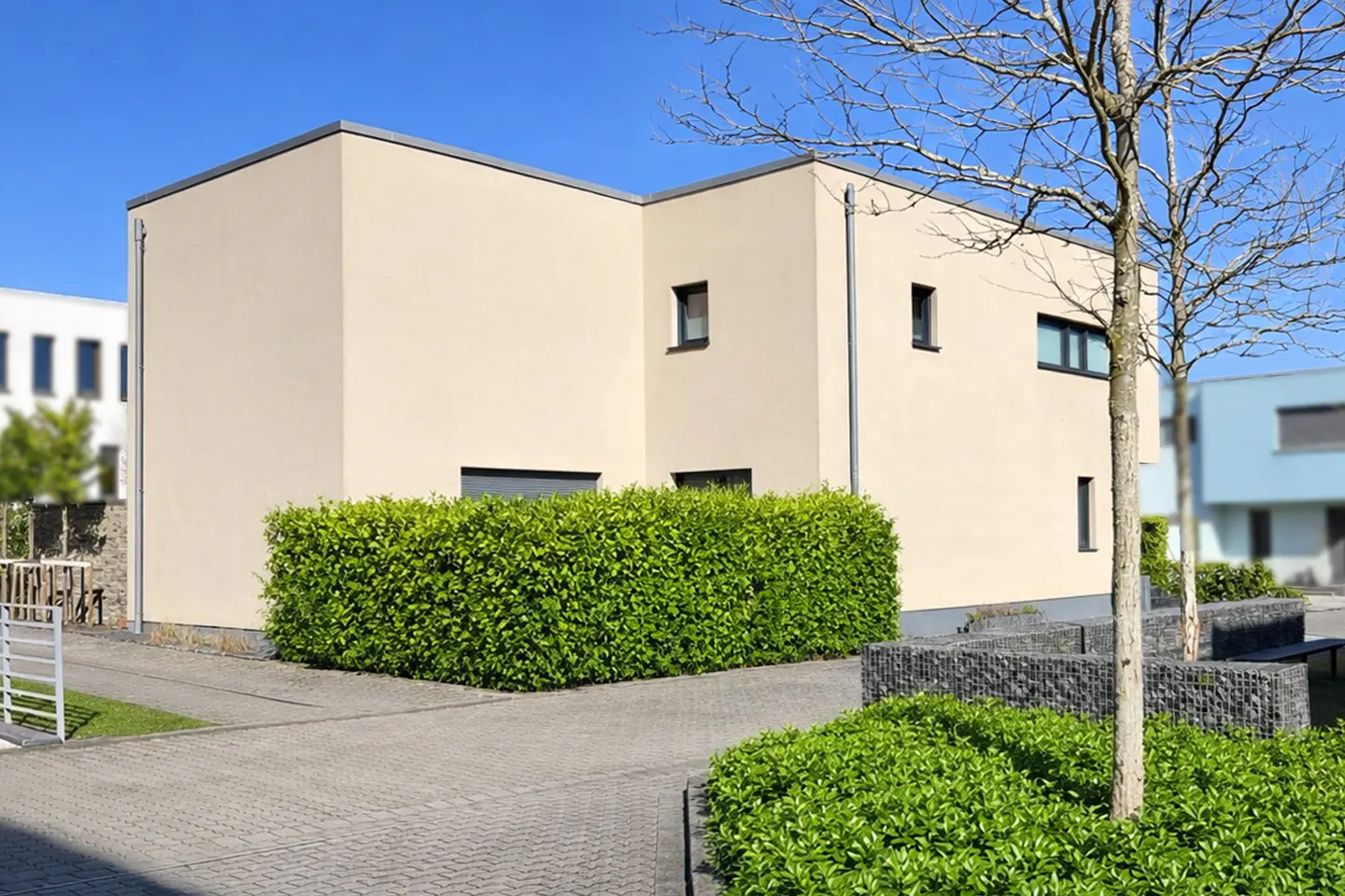 Modern beige house with black framed windows, green hedge, and gray stone walkway under a clear blue sky.
