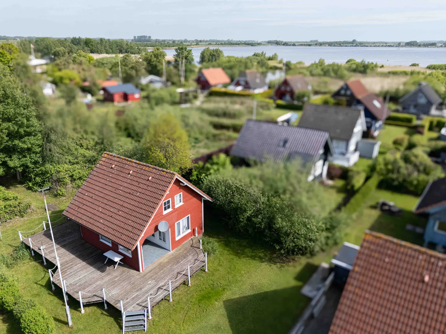 Aerial view of a red house with a brown roof and a wooden deck in a green, lakeside neighborhood.
