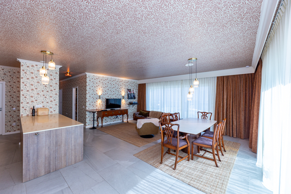 Open-plan living area with floral wallpaper, a kitchen island, dining table with six chairs, and a cozy lounge space. [AI-generated text]