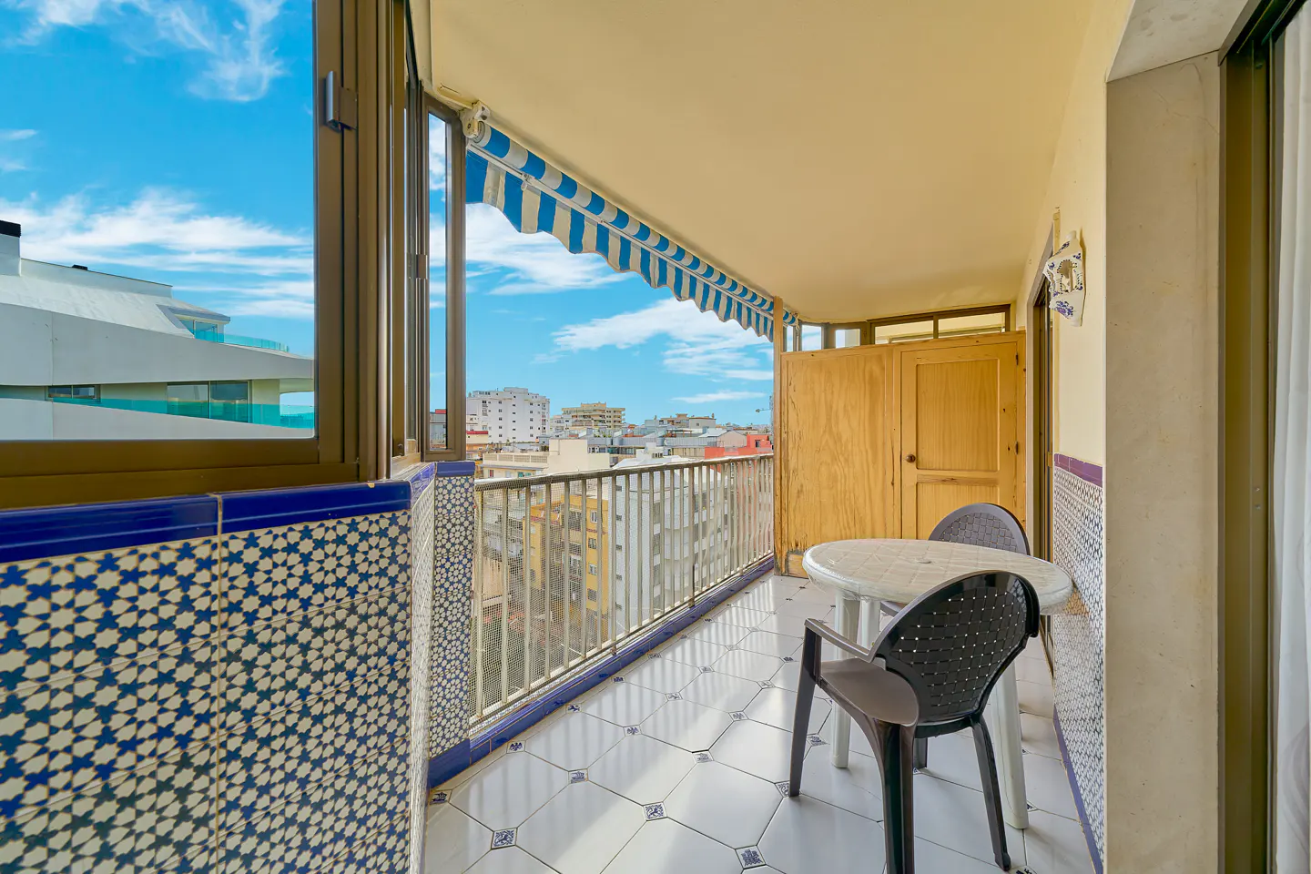 Balcony with blue and white patterned tiles, a small table, two chairs, and a blue-striped awning overlooking a city view.