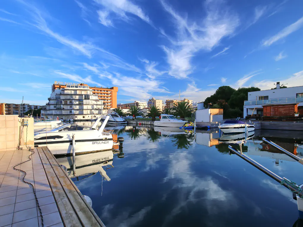 Marina view with boats docked in calm water, reflecting blue sky and clouds. Buildings and palm trees line the waterfront.