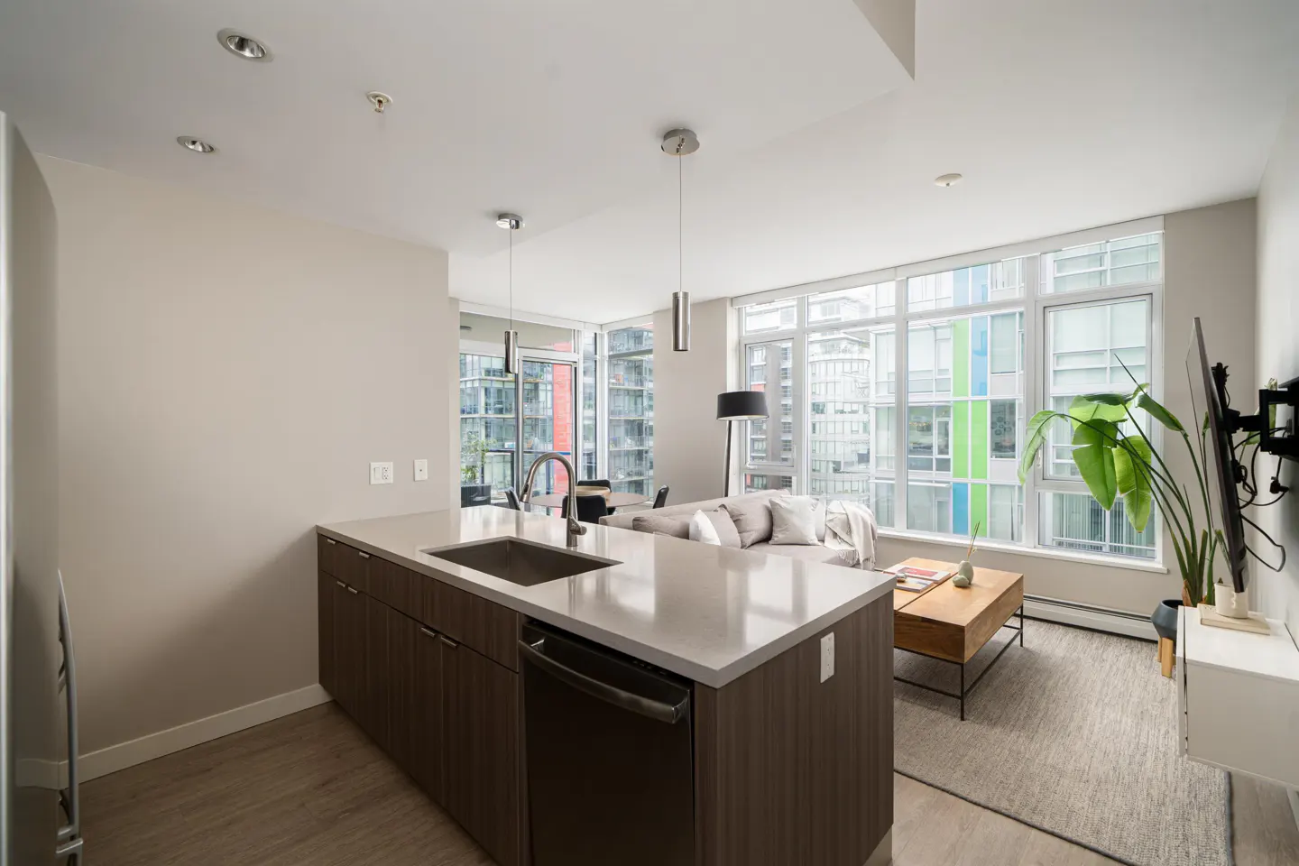 Bright, modern condo interior with a kitchen island, sink, and stainless steel appliances. Large windows overlook a city view.