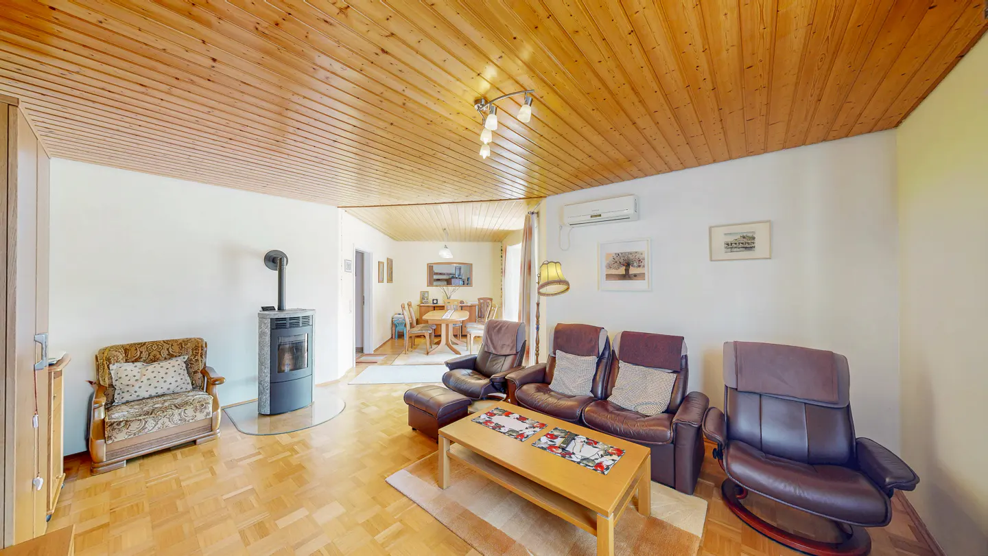 A living room with wood floors, a wood ceiling, and white walls. There is a wood-burning stove, brown leather furniture, and a wood coffee table.