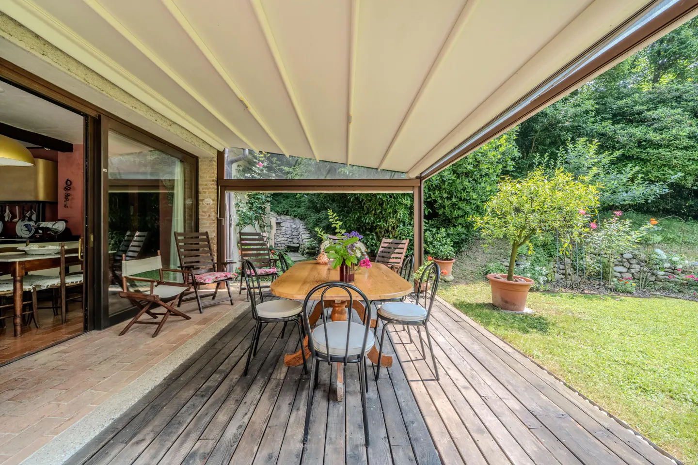 Covered patio with wood table, chairs, and garden view. The patio has a retractable awning and a wooden deck. The garden has green grass and trees.