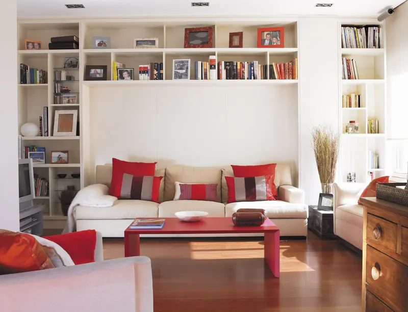 Living room with white built-in shelves filled with books and photos. A beige sofa with red pillows sits below, and a red coffee table is in front.