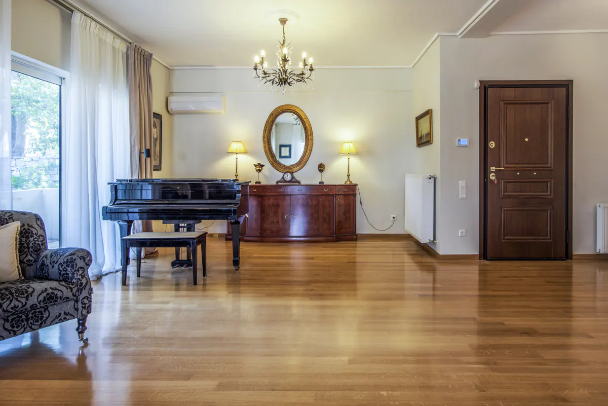 A living room with hardwood floors, a black piano, a brown wooden dresser with an oval mirror, and a chandelier.