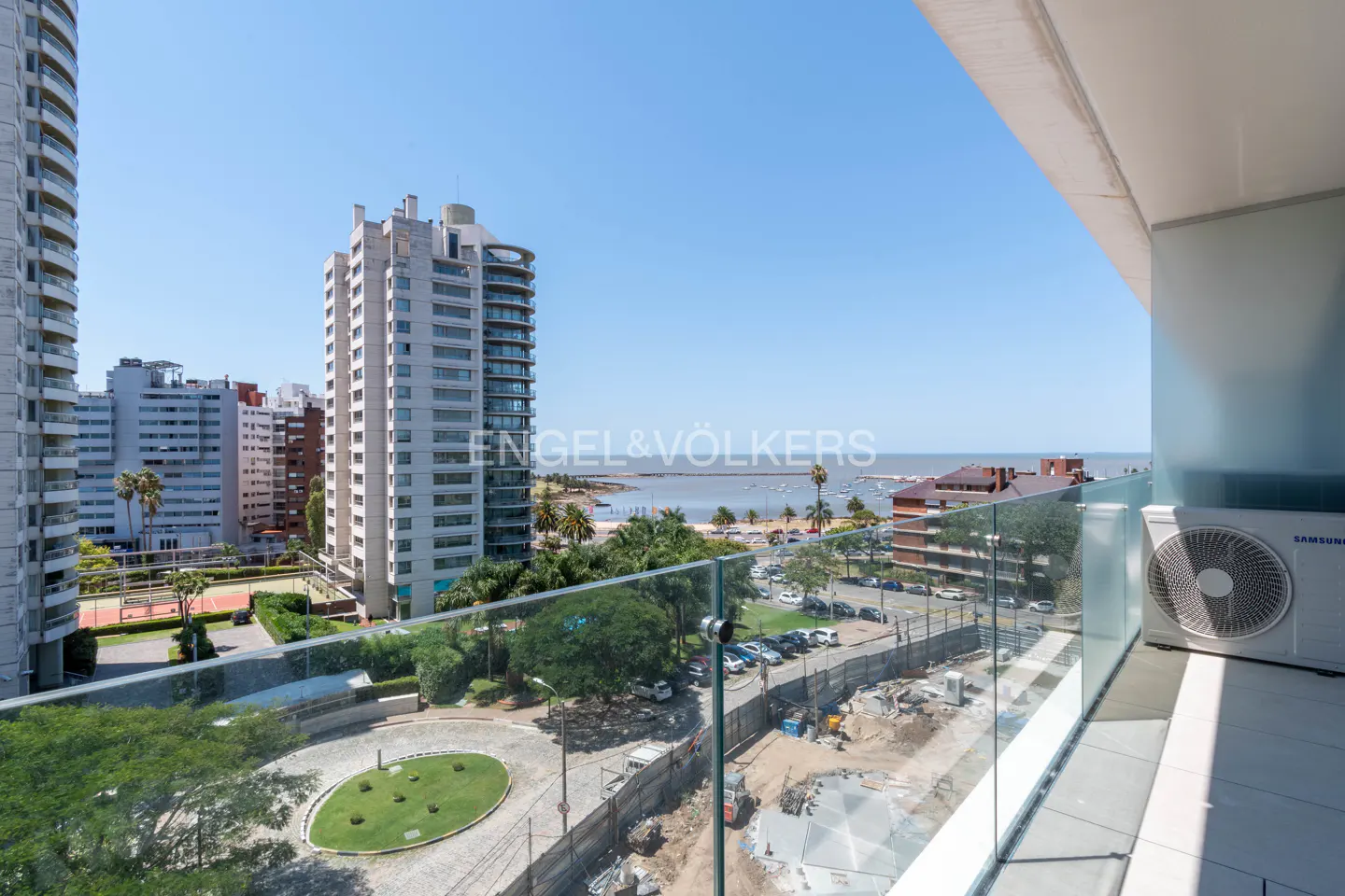 View from a balcony with glass railings overlooking a coastal city with tall buildings, a beach, and the ocean under a blue sky.