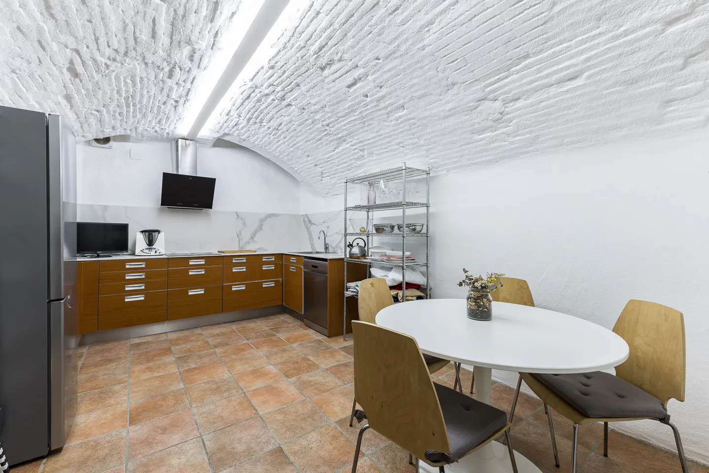 A kitchen with a white brick ceiling, brown cabinets, and a white table with four chairs. A stainless steel refrigerator is on the left.