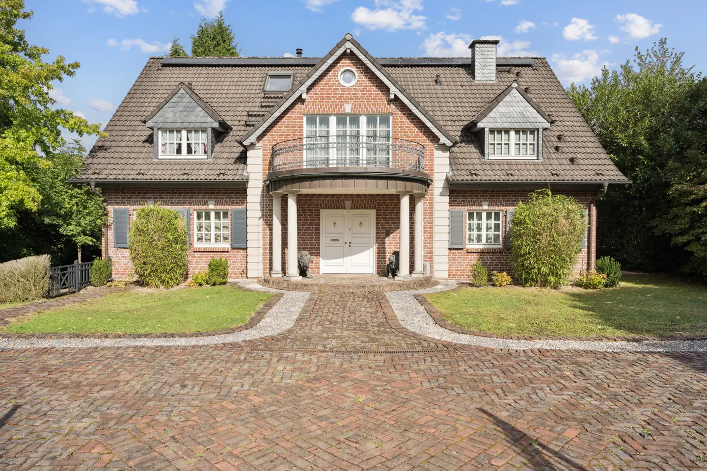 Brick house with a gray tiled roof, white trim, and a brick driveway. A balcony sits above the front door.