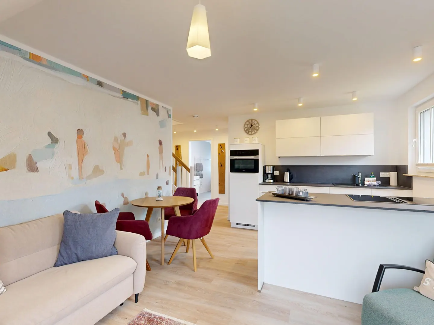 Open-concept living space with a beige sofa, round wood table with red chairs, and a white kitchen with black countertops.