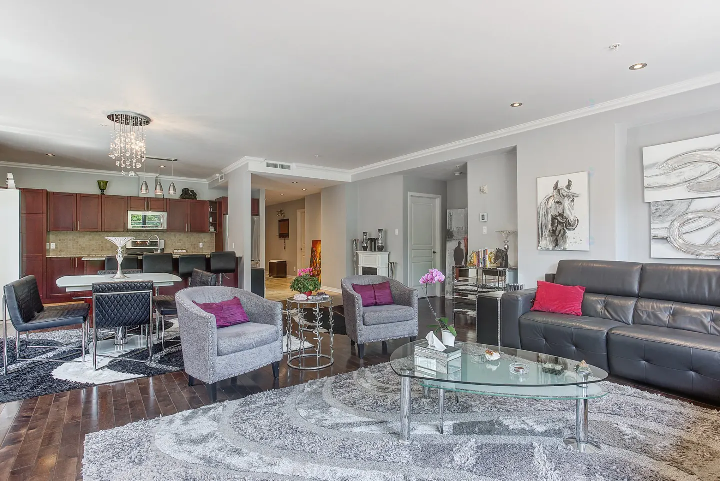 Open-concept living space with a black leather sofa, gray armchairs, and a glass coffee table on a gray patterned rug. Kitchen in the background.