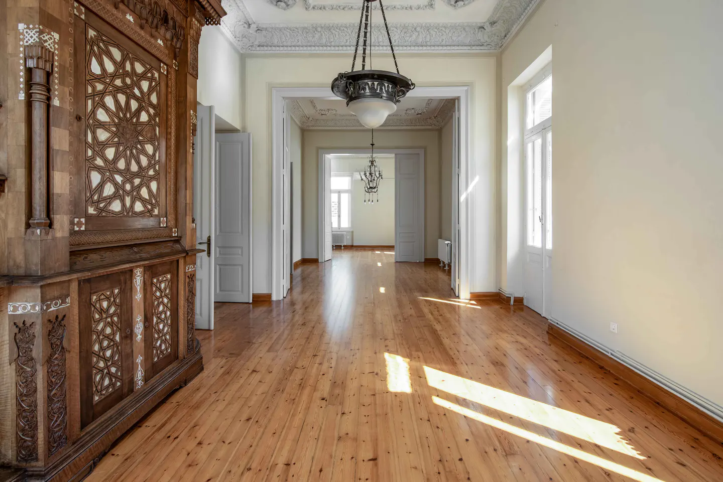 Hallway with wood floors, white walls, and a large, ornate wooden cabinet with geometric patterns. A chandelier hangs from the decorative ceiling.
