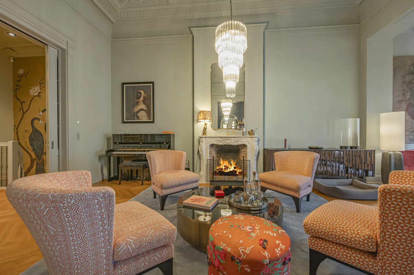 Living room with orange patterned chairs, fireplace, piano, and chandelier. Walls are light green.