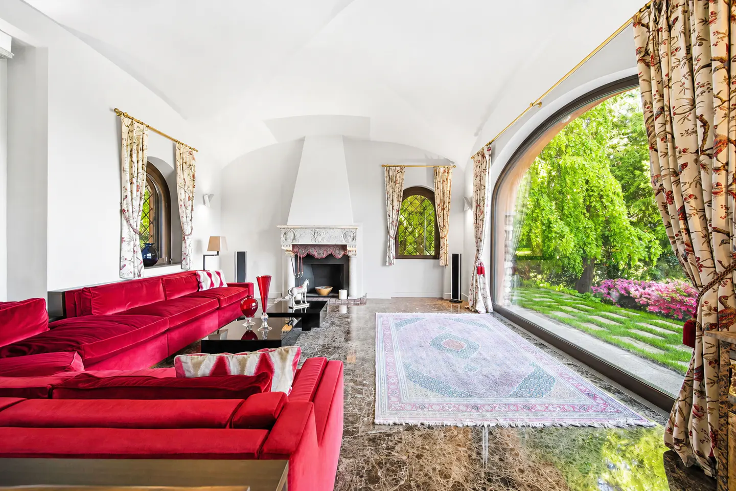 Bright living room with a large red sectional sofa, marble floors, a fireplace, and a wall of windows overlooking a garden.