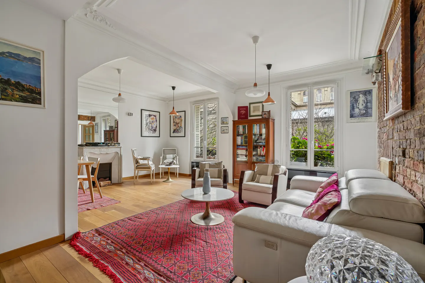 Bright living room with white walls, wood floors, and a red patterned rug. A white sofa faces a brick wall with art. Windows overlook a garden.