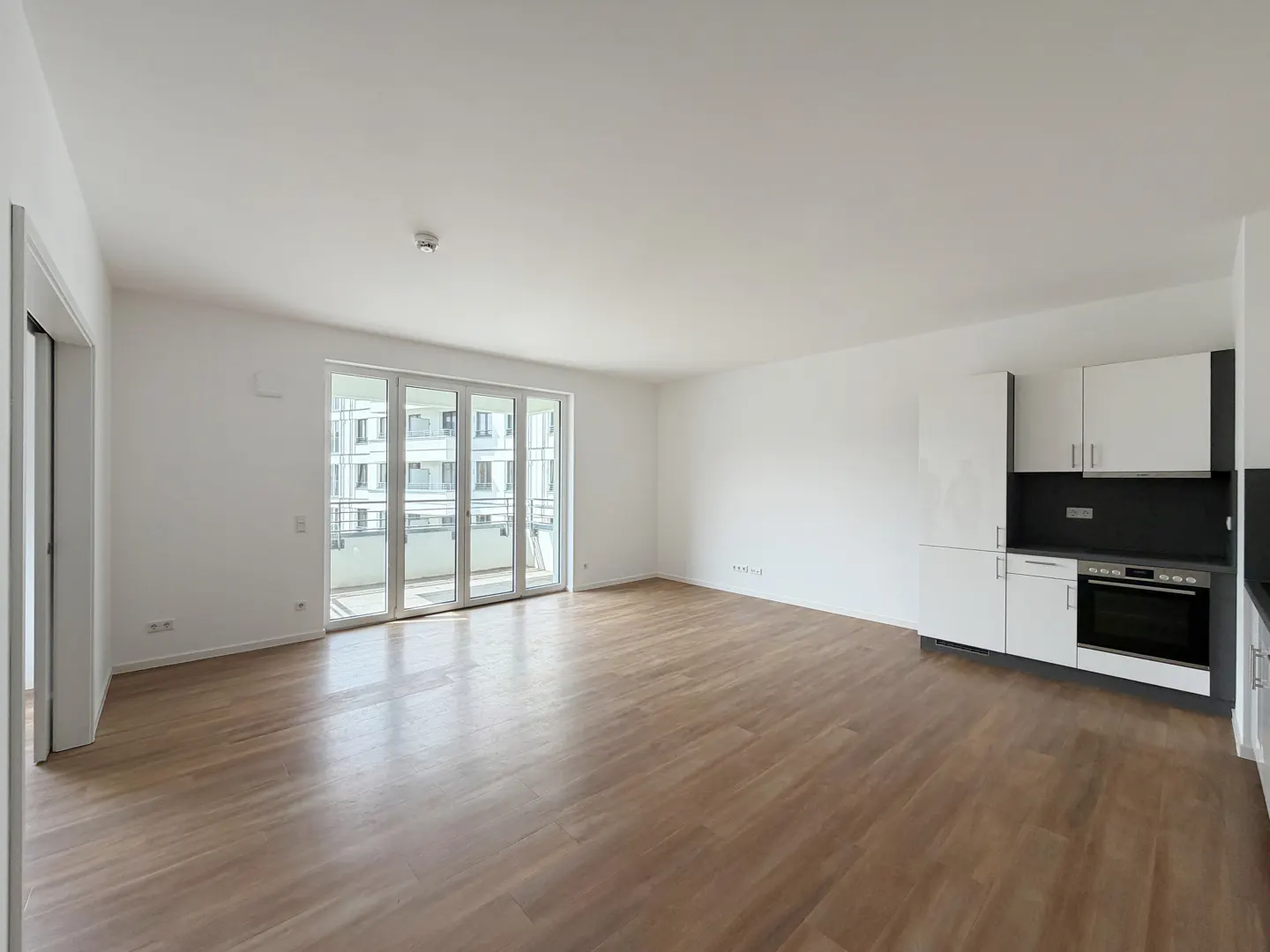 Bright, empty apartment with wood floors, white walls, and a kitchen area with white cabinets. Balcony doors offer a view of another building.