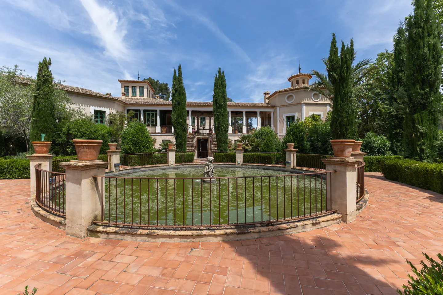 A circular fountain with a metal fence sits on a terracotta patio before a Mediterranean-style villa with tall cypresses.