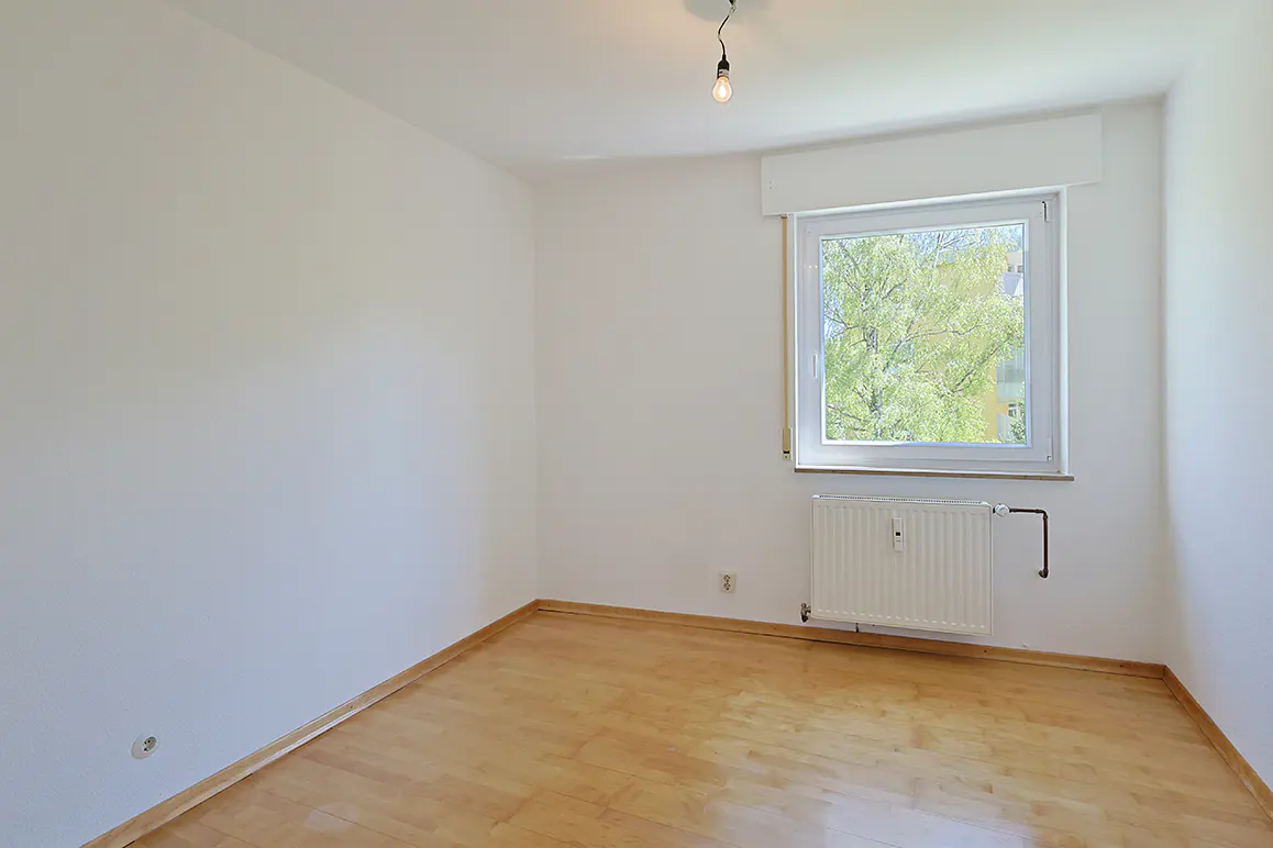 Empty room with white walls, light wood flooring, a single window with a view of trees, and a white wall-mounted radiator.