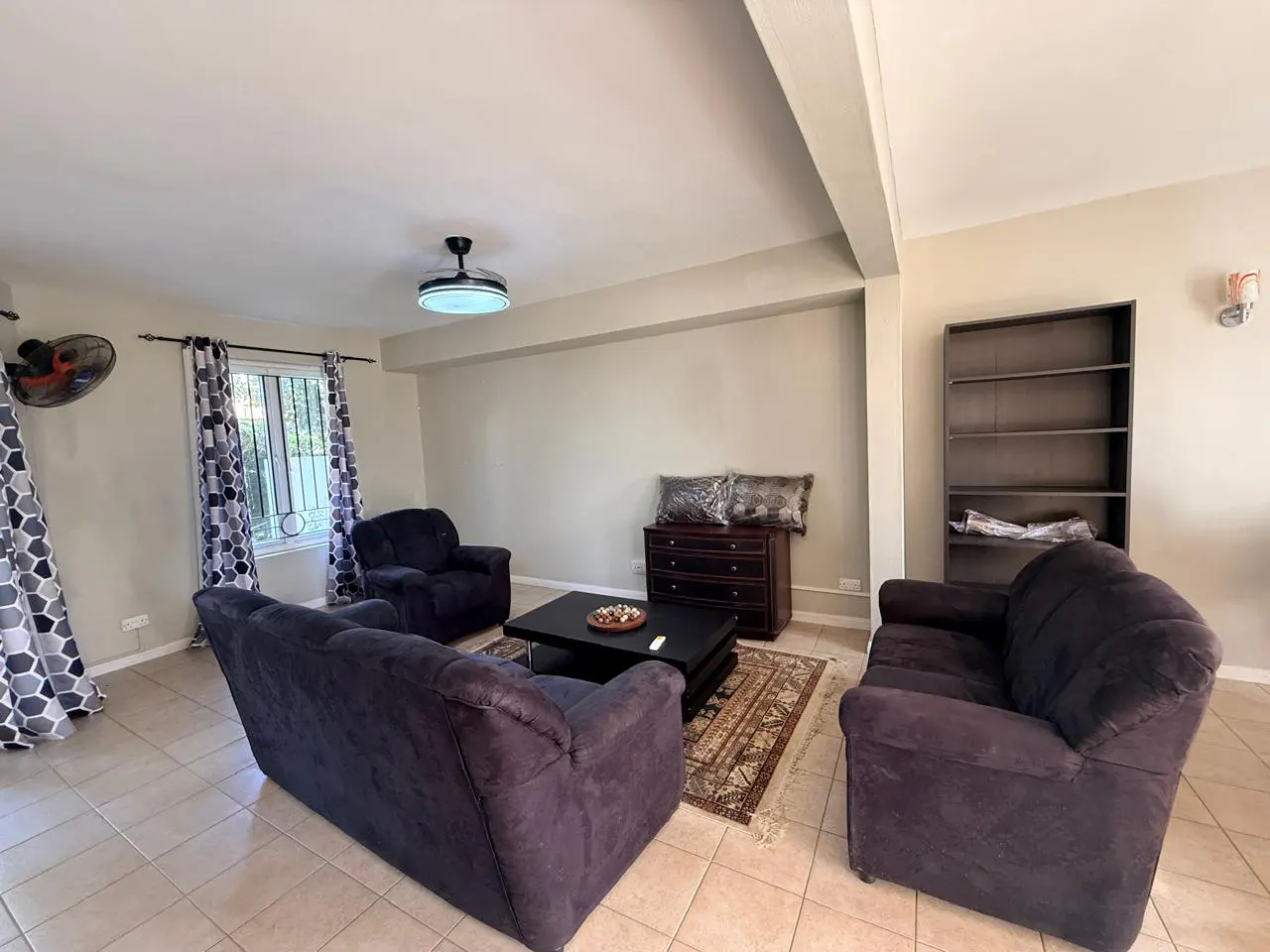 Living room with tile floor, dark sofas, black coffee table, and dark wood dresser and bookshelf. Curtains frame a window.